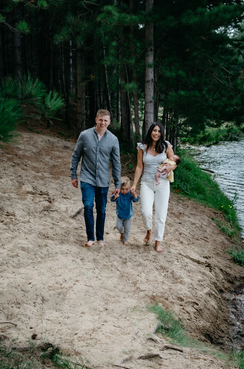 Family photo of Dad, mom, brother and newborn baby walking next to a lake with pine trees in the background