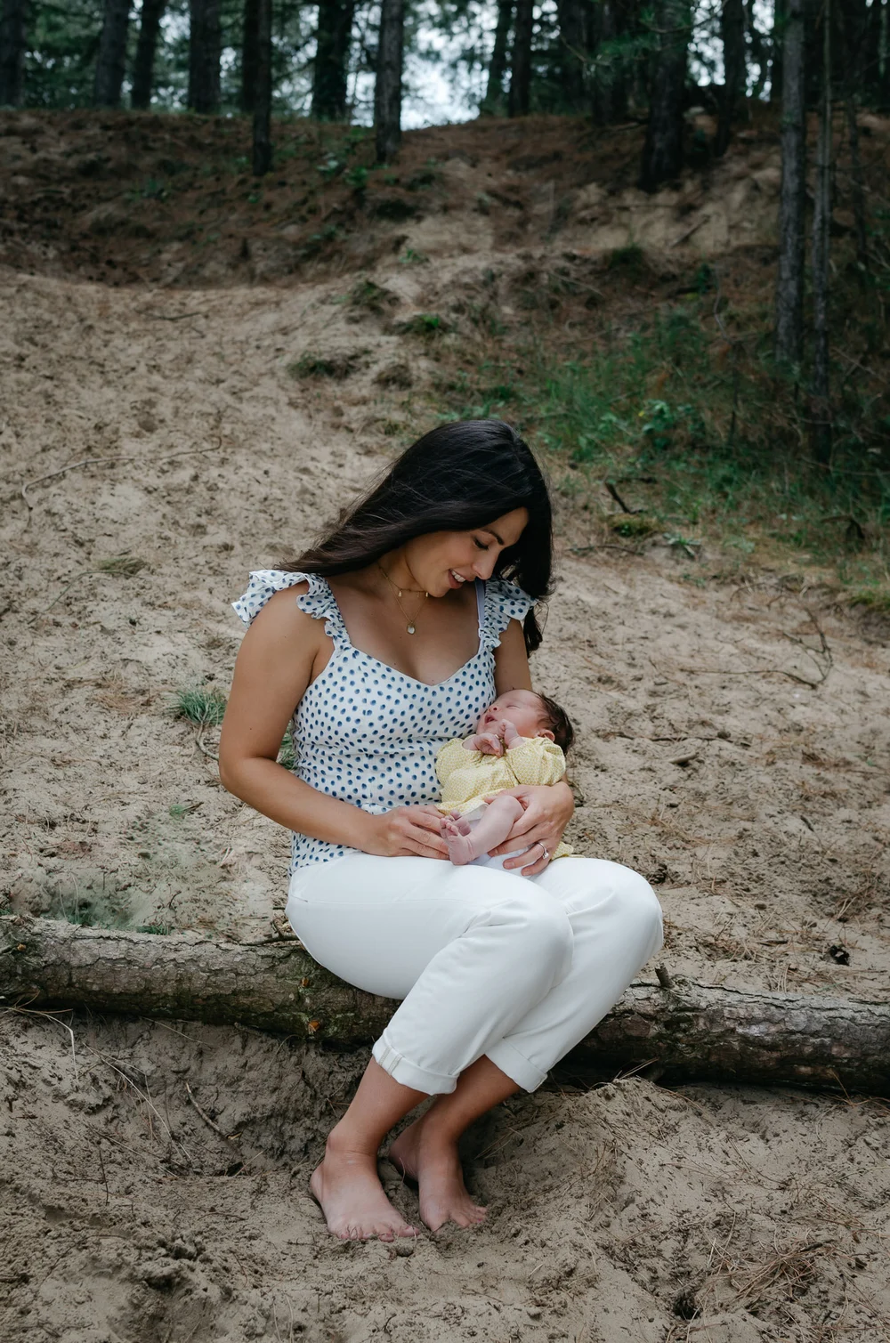 Mom holding her newborn baby while sitting on a piece of wood on a beach with pine trees in the background