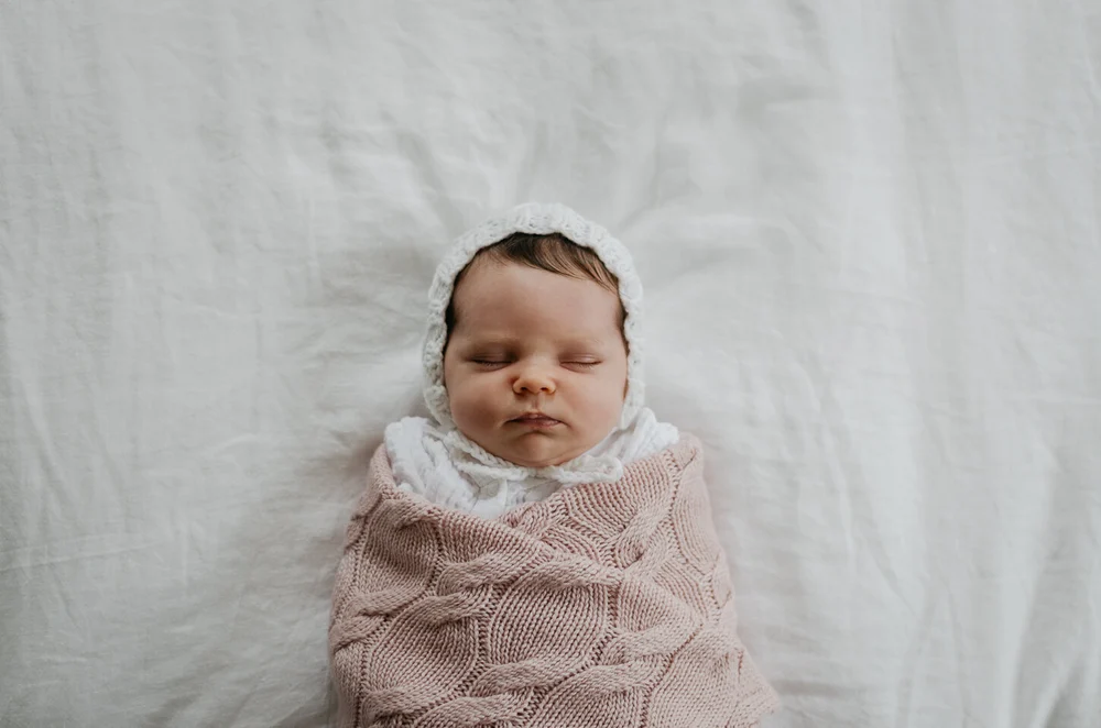 Newborn baby wrapped in a pink knitted blanket on a white bed, baby has a bonnet on her head.