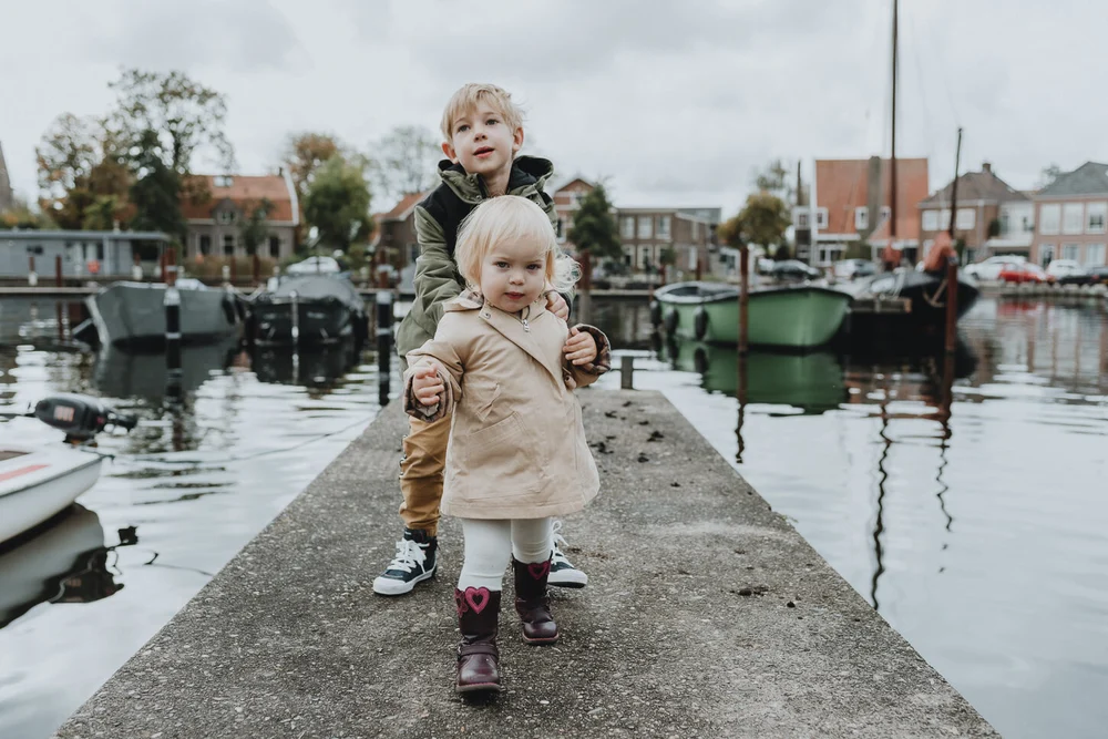 Brother and sister holding on to each other while standing on a dock with boats and Dutch houses in the background
