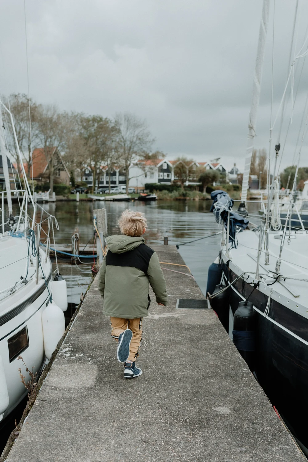 Boy running on a dock next to boats, the sky is grey and dramatic, the image was taken during a family photoshoot in Spaarndam