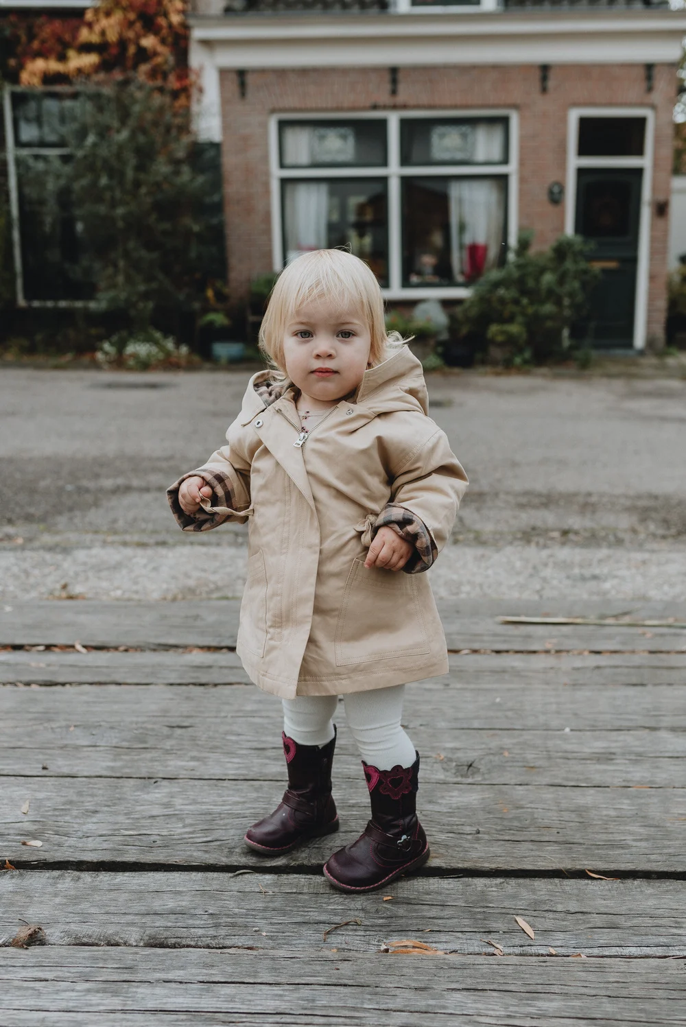 Cute little Dutch girl wearing her light brown coat and brown boots standing on a wood bridge with a cute Dutch house in the background