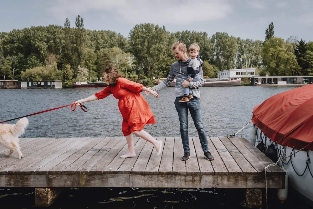 Family standing on a dock next to the water while dad is holding the toddler and mom is chasing after the dog, very fun photo